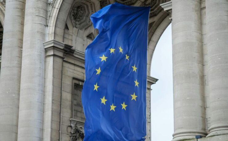 Duża flaga Unii Europejskiej z żółtymi gwiazdami na niebieskim tle jest wyświetlana między wysokimi kamiennymi kolumnami zabytkowego budynku, symbolizując jedność i wspólne standardy, takie jak system EORI w krajach członkowskich.