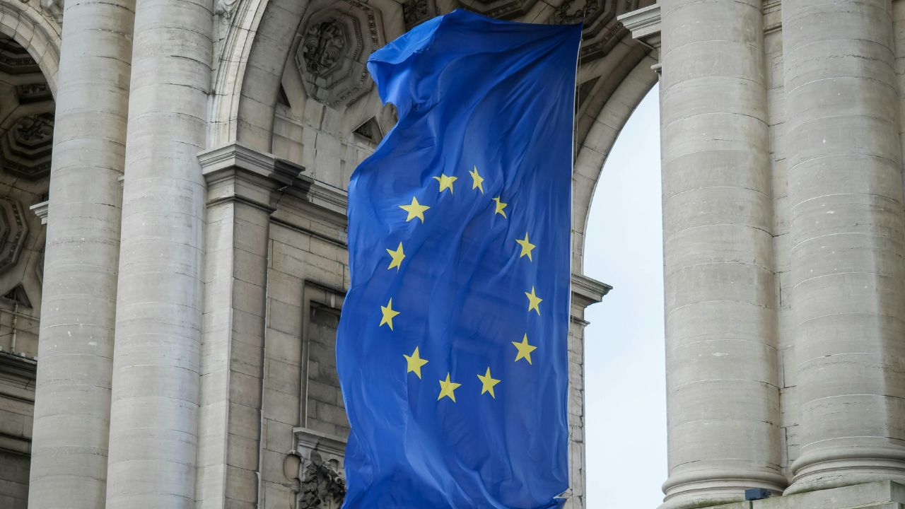 Duża flaga Unii Europejskiej z żółtymi gwiazdami na niebieskim tle jest wyświetlana między wysokimi kamiennymi kolumnami zabytkowego budynku, symbolizując jedność i wspólne standardy, takie jak system EORI w krajach członkowskich.