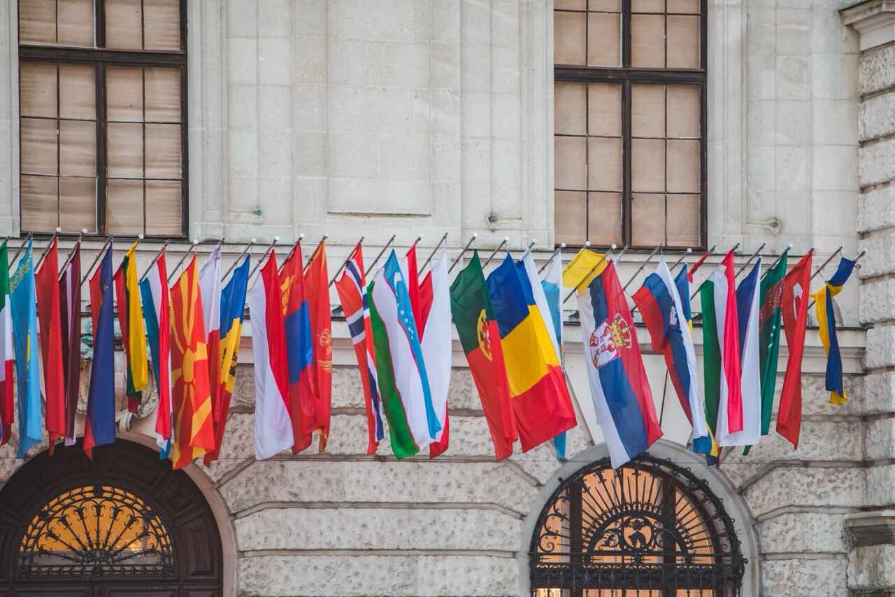 Rząd różnych flag narodowych z różnych krajów, symbolizujących międzynarodową współpracę Intrastat, jest wyświetlany przed zabytkowym budynkiem z łukowatymi drzwiami i dużymi oknami.