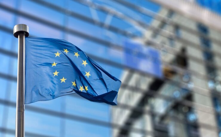 Flaga Unii Europejskiej z żółtymi gwiazdami na niebieskim tle faluje przed nowoczesnym szklanym budynkiem, którego okna odzwierciedlają flagę - odpowiednia scena do dyskusji na temat Intrastat i statystyk handlu transgranicznego.