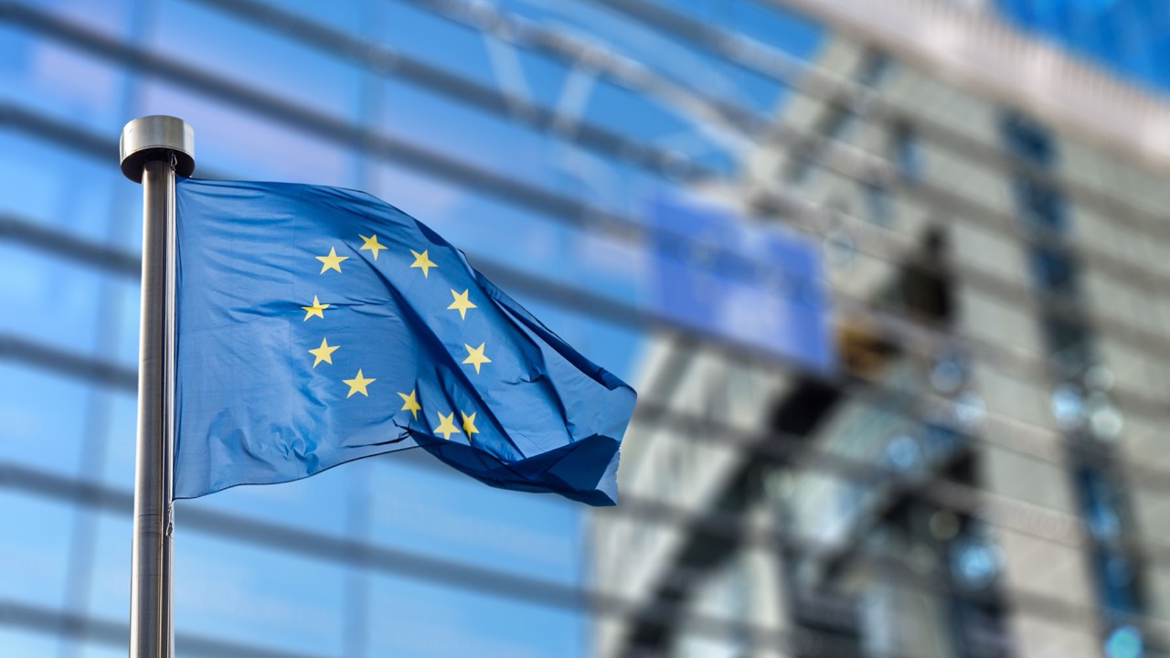 Flaga Unii Europejskiej z żółtymi gwiazdami na niebieskim tle faluje przed nowoczesnym szklanym budynkiem, którego okna odzwierciedlają flagę - odpowiednia scena do dyskusji na temat Intrastat i statystyk handlu transgranicznego.