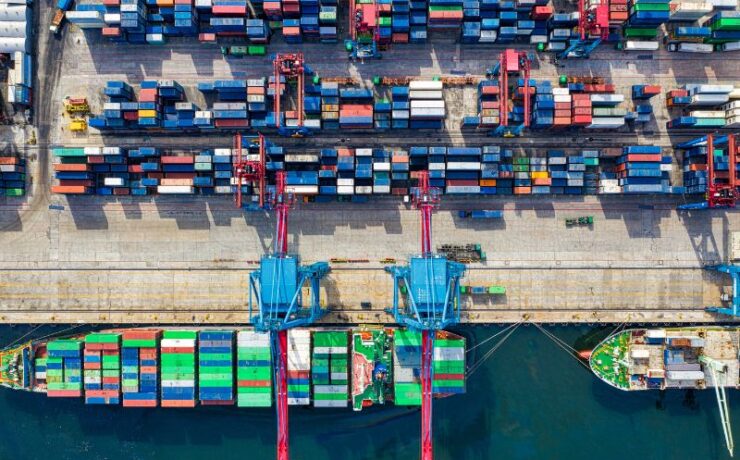 Widok z lotu ptaka na ruchliwy port żeglugowy z zacumowanymi dwoma dużymi statkami towarowymi, kolorowymi kontenerami ułożonymi na lądzie i statkach oraz dźwigami przenoszącymi kontenery - podkreślając wydajny proces odprawa importowa w tętniącym życiem terminalu.