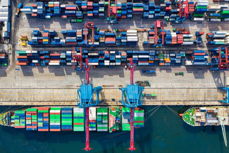 Widok z lotu ptaka na ruchliwy port żeglugowy z zacumowanymi dwoma dużymi statkami towarowymi, kolorowymi kontenerami ułożonymi na lądzie i statkach oraz dźwigami przenoszącymi kontenery - podkreślając wydajny proces odprawa importowa w tętniącym życiem terminalu.