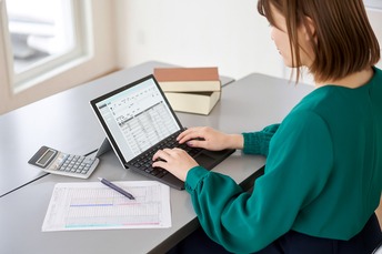 Kobieta w zielonej bluzce pracuje na laptopie wyświetlającym arkusz kalkulacyjny w Agencji Celnej Kraków. Obok niej na stole leży kalkulator, wydrukowany arkusz kalkulacyjny, długopis i dwie zamknięte książki.