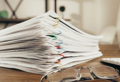 Dokumenty-celne Stos papierów, prawdopodobnie dokumentów celnych, spiętych kolorowymi spinaczami, leży na drewnianym biurku. Na pierwszym planie para okularów spoczywa obok stosu, podczas gdy półki i niewyraźne obiekty pojawiają się w tle.
