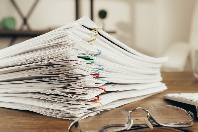 Stos papierów, prawdopodobnie dokumentów celnych, spiętych kolorowymi spinaczami, leży na drewnianym biurku. Na pierwszym planie para okularów spoczywa obok stosu, podczas gdy półki i niewyraźne obiekty pojawiają się w tle.