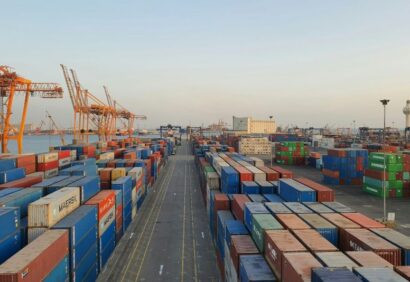 Eksportowanie-towaru-poza-UE Duży port żeglugowy z rzędami ułożonych w stos kontenerów w różnych kolorach i kilkoma wysokimi pomarańczowymi dźwigami, pod czystym niebem, z wodą i budynkami przemysłowymi w tle - idealny do eksportu towarów poza UE.