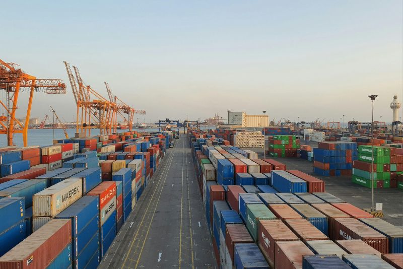 Duży port żeglugowy z rzędami ułożonych w stos kontenerów w różnych kolorach i kilkoma wysokimi pomarańczowymi dźwigami, pod czystym niebem, z wodą i budynkami przemysłowymi w tle - idealny do eksportu towarów poza UE.