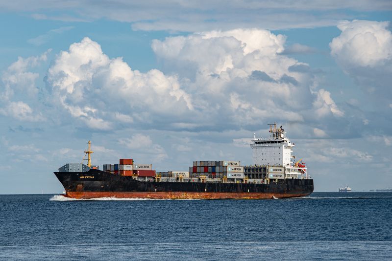 Duży kontenerowiec podróżuje przez ocean pod częściowo zachmurzonym niebem, przewożąc na swoim pokładzie ułożone w stosy kontenery transportowe w ramach eksportu towarów.