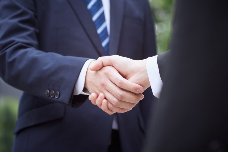 Dwie osoby w garniturach podają sobie ręce, sugerując profesjonalne porozumienie lub spotkanie, prawdopodobnie dotyczące braku odprawy celnej. Widoczne są tylko ich dłonie i część garniturów, z rozmytym tłem.