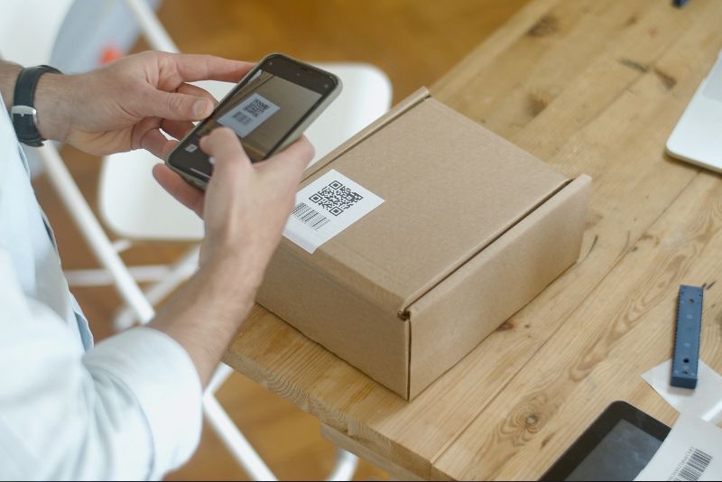 Osoba trzyma smartfon i skanuje kod QR, prawdopodobnie zawierający kod celny, na brązowym kartonowym opakowaniu umieszczonym na drewnianym stole. W pobliżu znajduje się białe krzesło, linijka i część laptopa.