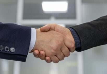 agencja-celna-czym-sie-zajmuje-2 Dwie osoby w garniturach podają sobie ręce, symbolizując porozumienie lub partnerstwo - być może finalizując szczegóły związane ze współpracą z agencją celną - na rozmytym tle biura.