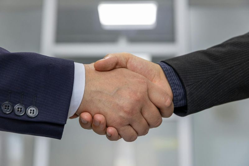 Dwie osoby w garniturach podają sobie ręce, symbolizując porozumienie lub partnerstwo - być może finalizując szczegóły związane ze współpracą z agencją celną - na rozmytym tle biura.