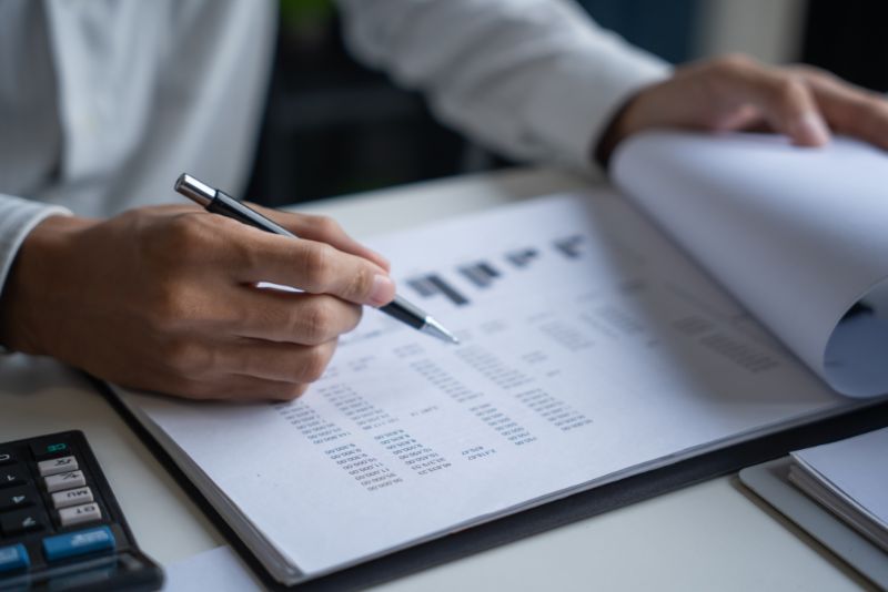 Osoba w białej koszuli przegląda dokumenty finansowe przy biurku, prawdopodobnie pracując dla agencji celnej. Trzyma długopis przerzucając papiery. W pobliżu widoczny jest kalkulator.