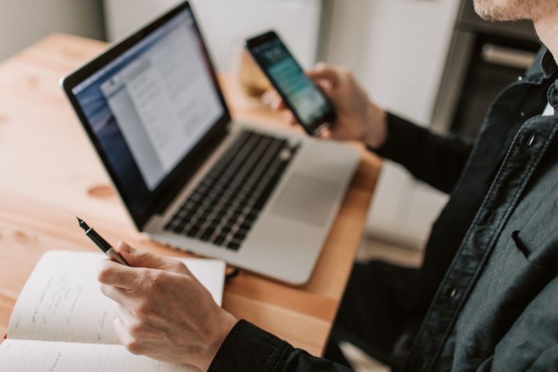 Mężczyzna trzyma w ręku telefon, długopis i wykonuje czynności na laptopie.