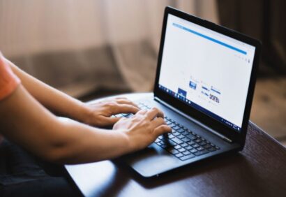 samodzielna-odprawa-celna Osoba pisze na czarnym laptopie umieszczonym na drewnianym stole. Na ekranie wyświetlana jest strona internetowa związana z eksportem towarów poza UE, z widocznymi kilkoma sekcjami, choć szczegóły są rozmyte i nieczytelne.