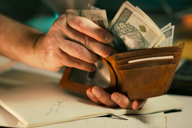 Osoba trzyma brązowy portfel wypełniony banknotami polskiej waluty, wyjmując gotówkę - być może w celu pokrycia kosztów odprawy celnej - siedząc przy biurku z otwartym notatnikiem i długopisem.