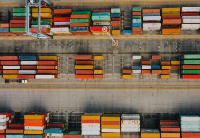 odprawa-celna-co-to Widok z lotu ptaka na port morski z rzędami kolorowych kontenerów towarowych ustawionych wzdłuż doków, dźwigiem ustawionym między nimi i ciężarówką widoczną w pobliżu kontenerów. Pokazane towary czekają na zatwierdzenie odprawy celnej.
