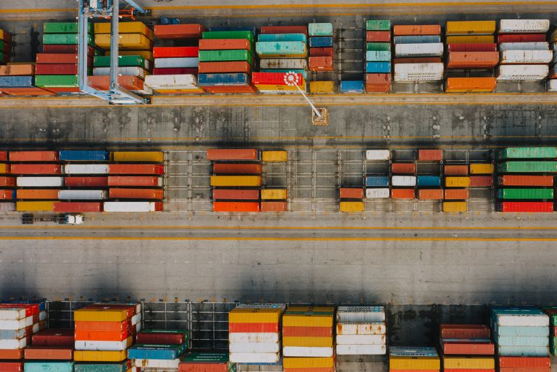 Widok z lotu ptaka na port morski z rzędami kolorowych kontenerów towarowych ustawionych wzdłuż doków, dźwigiem ustawionym między nimi i ciężarówką widoczną w pobliżu kontenerów. Pokazane towary czekają na zatwierdzenie odprawy celnej.