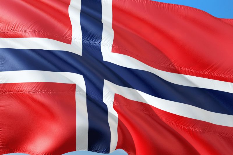 Obraz przedstawia flagę Norwegii, która ma czerwone tło z niebieskim krzyżem obrysowanym na biało, falującym na tle czystego, błękitnego nieba.