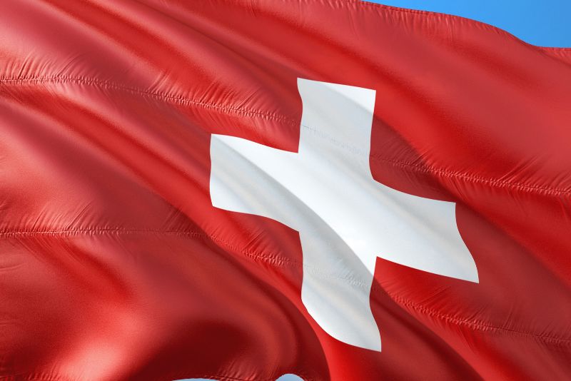 Czerwona flaga z białym krzyżem pośrodku faluje na wietrze na tle błękitnego nieba. Jest to flaga narodowa Szwajcarii, często widywana przez firmy, które organizują eksport do Szwajcarii.