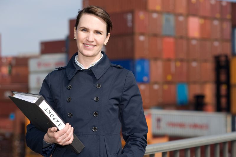 Kobieta w ciemnym płaszczu trzyma folder z napisem EXPORT i uśmiecha się do kamery, stojąc na zewnątrz przed dużym stosem kolorowych kontenerów transportowych oznaczonych.