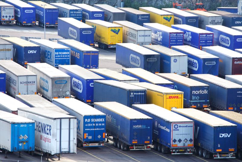 Widok z lotu ptaka na liczne zaparkowane ciężarówki i przyczepy towarowe z różnymi logo i kolorami firmy, ułożone w równoległych rzędach i ciasno upakowane razem.