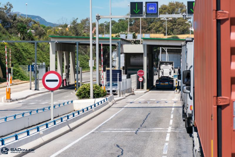 Ciężarówki i samochody osobowe ustawiają się w kolejce przy punkcie granicznym. Widać znaki stopu, szlabany i budki kontrolne; nad pasami ruchu widnieją zielone strzałki oraz znaki zakazu wjazdu.